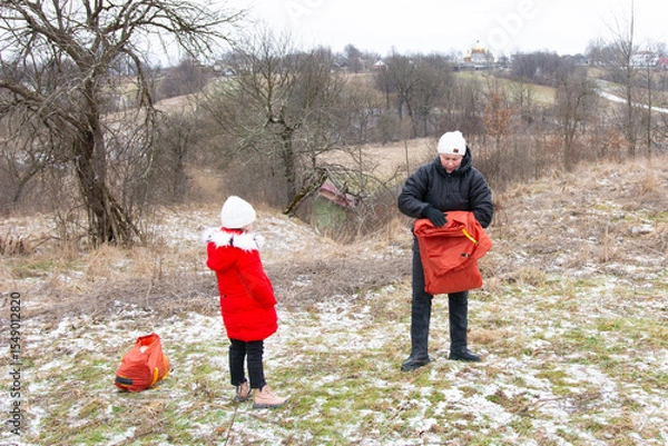 Fototapeta Bright winter adventure unfolds with a child and adult preparing gear in a snowy landscape surrounded by trees and distant hills