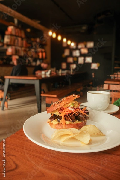 Fototapeta Close-up of gourmet sandwich bagel with shredded veggie mushrooms and chips served on white plate in cozy café setting with warm lighting and blurred background of diners and decor. Asunción, Paraguay