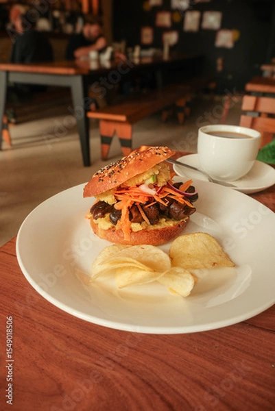 Fototapeta Close-up high angle shot of gourmet vegetarian sandwich bagel with mushrooms; chips served on white plate in cozy café setting. Coffee on table in background. Travel photography in Asunción, Paraguay