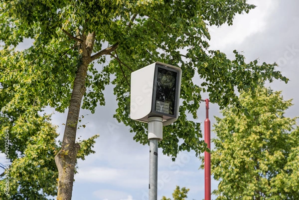Obraz A modern white surveillance speed camera (flitspaal) mounted on a pole amidst green trees and a bright sky, ensuring safety and monitoring.