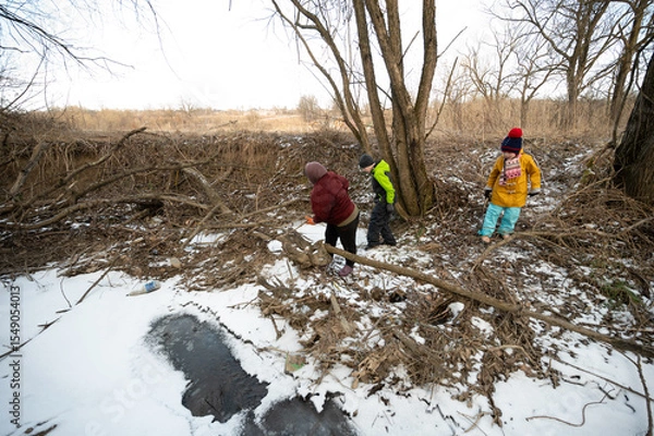 Fototapeta Children exploring a winter landscape while collecting sticks and playing near a frozen creek in a rural area during a sunny afternoon