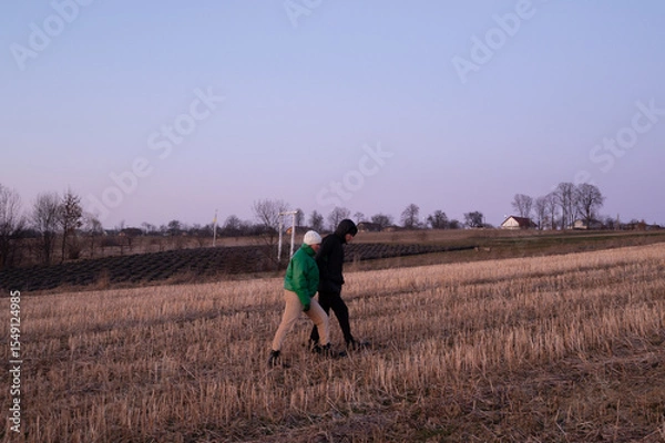 Fototapeta Two friends enjoy an evening stroll across the serene landscape of a rural field as the sky transitions into twilight during early spring