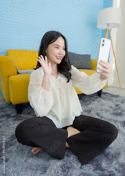Obraz Asian young beautiful happy woman wears casual clothes smile holding using mobile phone, smartphone media, using apps shopping online reading news sitting on floor in light blue living room at home.