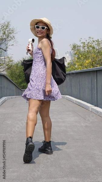 Obraz Young Asian woman stands on a paved walkway in a confident and relaxed pose dressed in a stylish, knee-length purple dress holding a portable fan