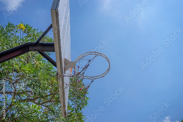 Fototapeta An old outdoor basketball hoop with a weathered wooden backboard captured from below under a bright blue sky. The frayed net and rustic look evoke nostalgia and street basketball vibes.