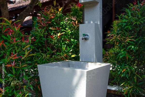 Fototapeta An outdoor sink with a metal faucet surrounded by green and red ornamental plants. The modern stone structure is situated in a shaded garden area, evoking cleanliness and nature.