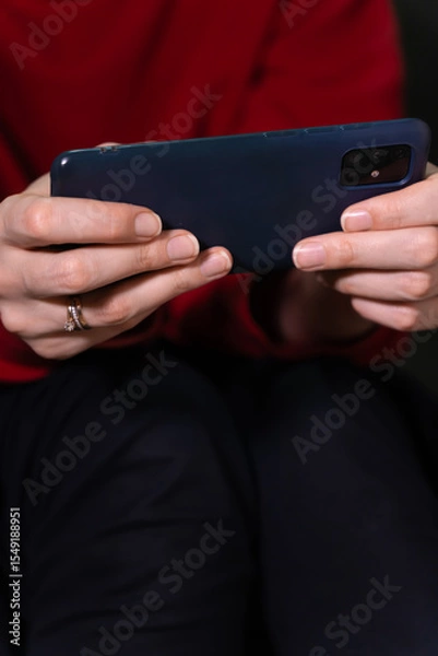 Obraz Woman wearing a vibrant red sweater and dark pants holds a smartphone horizontally in both hands, sitting on the toilet. Vertical photo