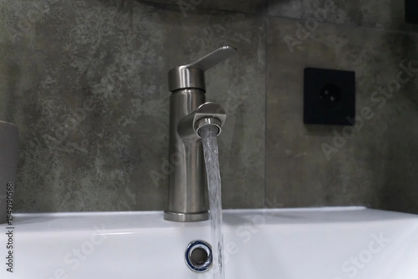 Obraz Modern bathroom faucet pouring water into sink with a gray tiled wall and black electric outlet in the background, promoting water conservation and modern bathroom design