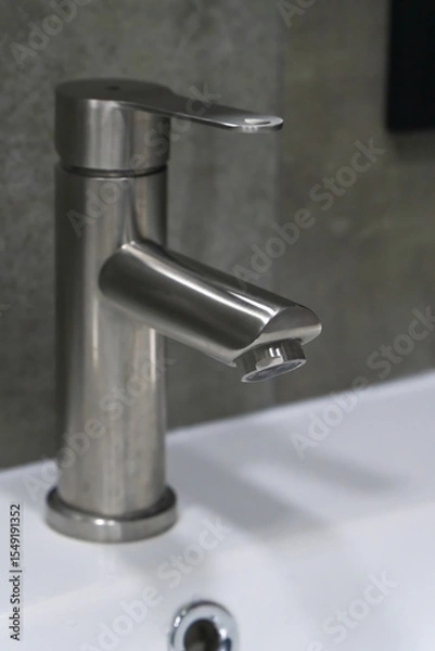 Obraz Close-up view of a sleek, modern stainless steel faucet positioned above a white ceramic sink, showcasing contemporary bathroom design and fixtures. Vertical photo