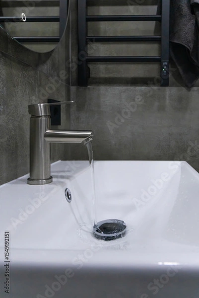 Obraz Close-up view of a modern bathroom sink featuring a stainless steel faucet, white ceramic basin, and a textured concrete wall, creating a stylish and minimalist atmosphere. Vertical photo