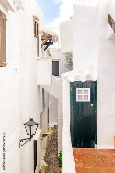 Fototapeta Charming Whitewashed Alley With Dark Green Door and Cobblestone Path in Binibeca Vell. Menorcan Architecture, Traditional Lantern, ideal for travel and design concepts