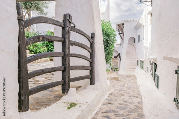 Fototapeta Traditional Wooden Gate and Whitewashed Architecture in Binibeca Vell. Menorcan Coastal Alley, Cobblestone Street, Mediterranean Village Charm