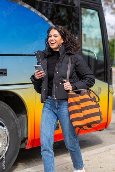 Obraz smiling woman walks beside a colorful bus while holding mobile phone