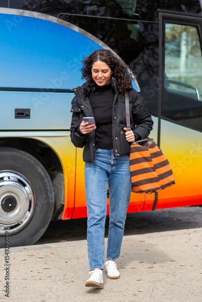 Obraz Full body smiling travel woman walking with bag while looking at her phone.