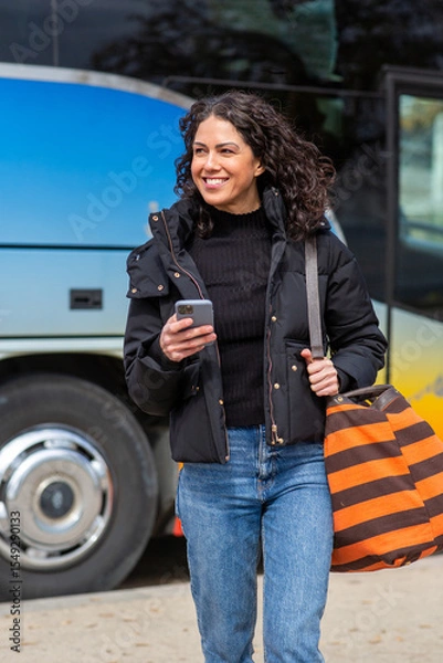 Obraz woman walking near colorful bus, smiling while holding her phone