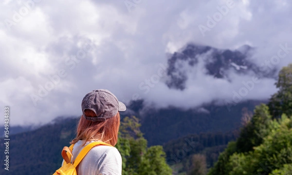 Fototapeta Young woman hiker in cap with orange backpack gazes at majestic mountains shrouded in clouds, surrounded by lush greenery, capturing the essence of adventure and exploration in nature