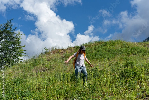 Fototapeta Young woman hiker in cap walking on a grassy hillside under a bright blue sky with fluffy clouds, enjoying nature and the outdoors while exploring the natural landscape
