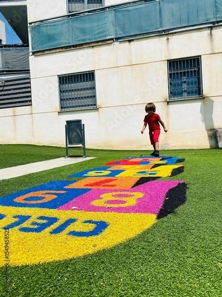Obraz Young boy in red clothes playing hopscotch on a vibrant play area in a sunny urban courtyard.