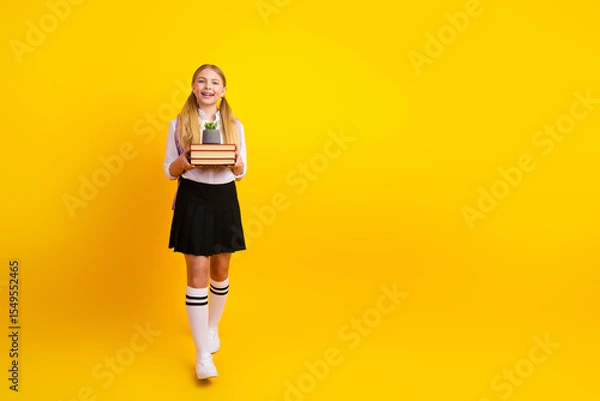 Obraz Cheerful schoolgirl standing against yellow background holding books and a small plant, representing education, joy, and learning