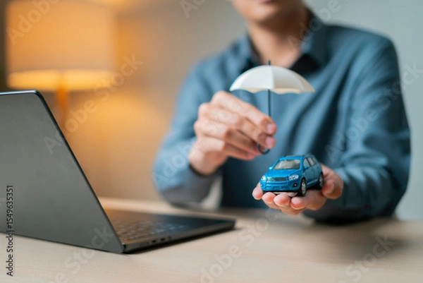 Fototapeta Hand holding a small blue toy car under a white umbrella beside a laptop, symbolizing online auto insurance protection.