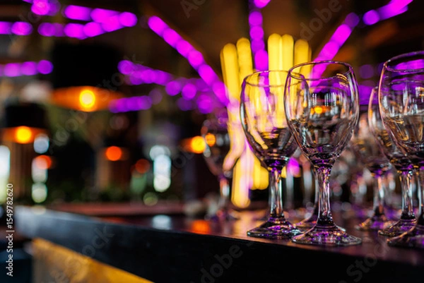 Fototapeta Empty clear wineglasses stand in rows on bar counter at nightclub, blurred purple neon and yellow lights on background. Clean cocktail glasses ready for celebrate party.