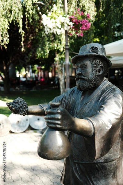 Obraz Bronze statue of a man offering grapes and a bottle, located in a park in town Vrsac, Serbia.