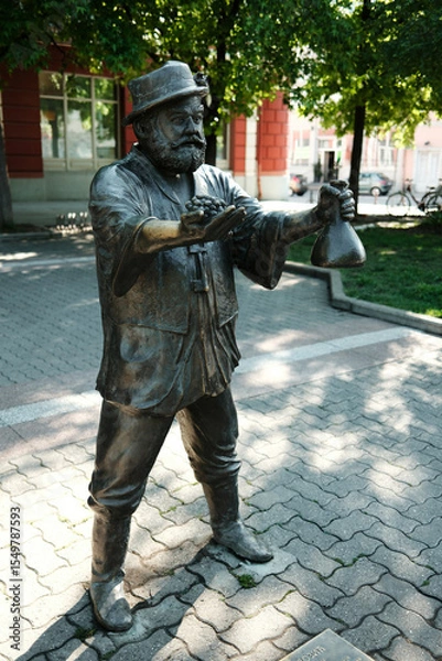 Obraz Full-body bronze sculpture of a man holding grapes and a wine bottle, standing in a paved park area in Vrsac, Serbia country.