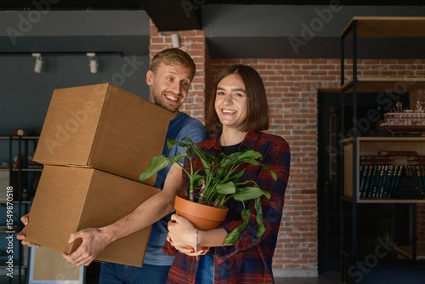Obraz A couple enjoying moving into a new home, carrying boxes and a plant.