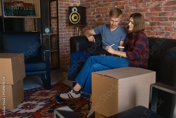 Obraz Couple on a couch planning with boxes around, in a modern cozy living space.