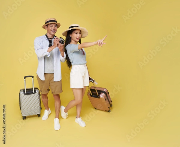 Fototapeta Happy Asian couple with hats luggage and camera pointing into distance Ready for adventure isolated on yellow background. Great for travel, holidays, or exploration themes.