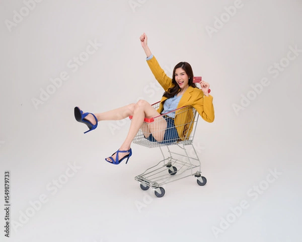 Fototapeta Ecstatic Asian woman in yellow blazer and heels holding a credit card joyfully sits in a shopping cart. Perfect for sales, promotions, or payment themes.
