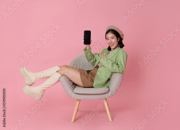 Fototapeta Happy Asian woman sitting in chair showing smartphone with blank white screen isolated on pink background. Ideal for app mockups or ad content.