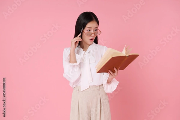 Fototapeta Young Asian woman surprised reading book with glasses and pen isolated on pink background. Expresses discovery amazement.