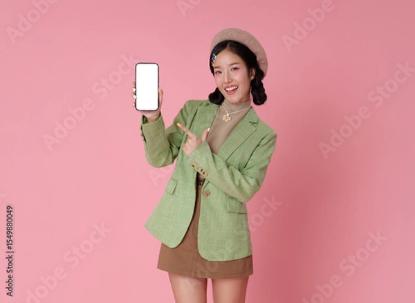Fototapeta Smiling Asian woman in green blazer and beret holds up phone with blank screen, pointing. isolated on Pink background, perfect for app promo or ad copy.