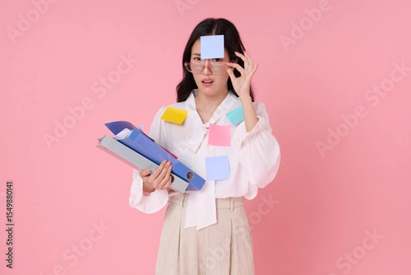 Fototapeta Asian woman feeling stressed with work overload, covered in colorful sticky notes and holding folders. Studio portrait on pink background, representing office pressure and multitasking.
