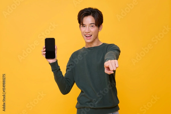 Fototapeta Happy Asian man showing smartphone mockup of blank screen and pointing finger isolated on yellow background.