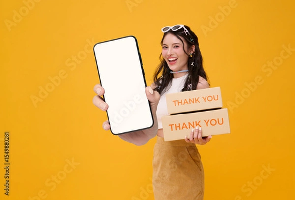 Fototapeta Happy young Asian woman smiling holding package parcel box and showing mobile phone application post mockup isolated on yellow background.
