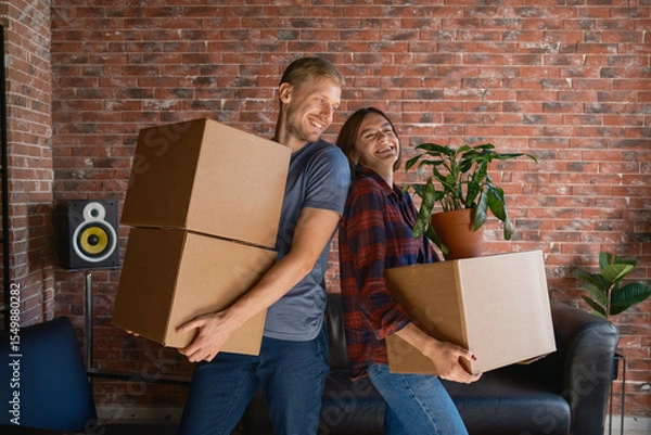 Obraz A joyful couple carries boxes and a plant, settling into their new residence