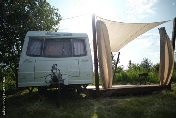 Fototapeta A camper van with a wooden terrace and fabric canopy is parked in the middle of tall grass and trees. This peaceful scene evokes a sense of freedom, glamping, and off-grid living.