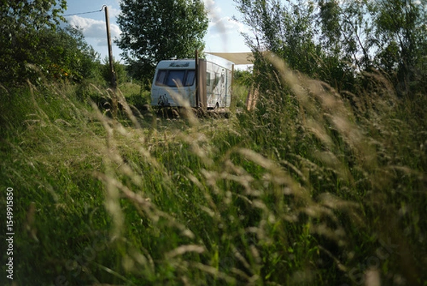 Fototapeta A camper van with a wooden terrace and fabric canopy is parked in the middle of tall grass and trees. This peaceful scene evokes a sense of freedom, glamping, and off-grid living.