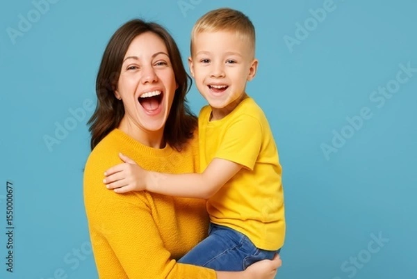 Obraz Smiling Mother in yellow sweater holds her happy son in yellow shirt and jeans in her arms. They both look at the viewer, laughing against a light blue background