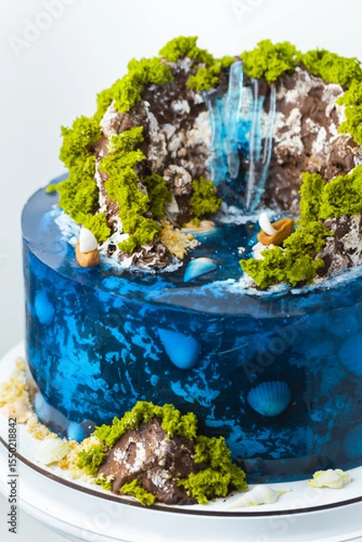 Fototapeta Beautiful birthday cake in the shape of an island decorated with chocolate edible mountains, greenery with waterfall. Tropical island cake surrounded with blue jelly ocean water