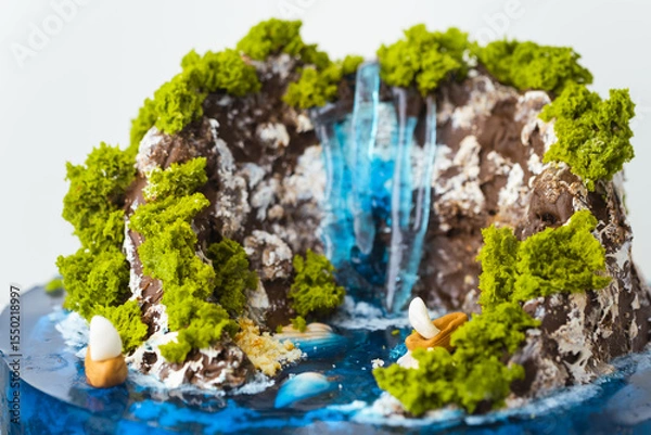 Fototapeta Beautiful birthday cake in the shape of an island decorated with chocolate edible mountains, greenery with waterfall. Tropical island cake surrounded with blue jelly ocean water