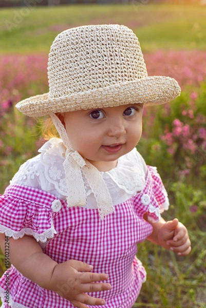 Obraz Beautiful one-year-old baby girl stands in a pink dress and a straw hat in a field at sunset among bright flowers.