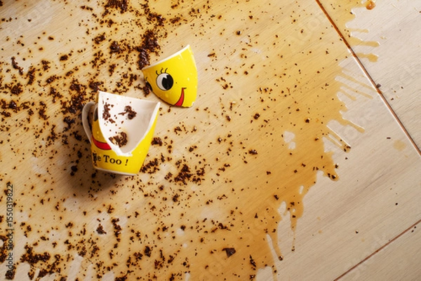 Fototapeta Broken yellow cup with smiley face and spilled black tea on the floor. Tea spilled on the floor. Splashes and tea leaves on the floor.