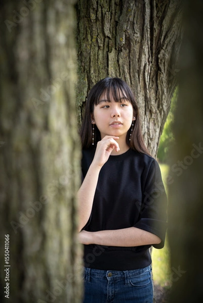 Fototapeta A young teenager stands pensively among tree trunks, framed by bark and soft natural light, wearing a black top and jeans in a calm forest scene.