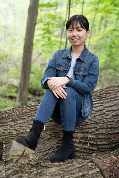 Fototapeta A young teenage girl in denim sits calmly on a fallen tree in a lush green forest, gazing off thoughtfully, surrounded by greenery.