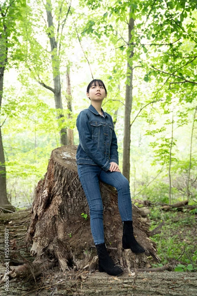 Fototapeta A young teenage girl in denim sits calmly on a fallen tree in a lush green forest, gazing off thoughtfully, surrounded by greenery.