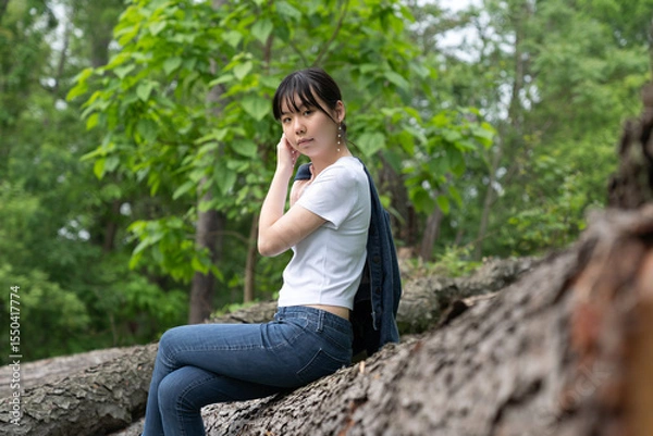 Fototapeta A young teenage girl in a white top and jeans sits calmly on a fallen tree in a lush green forest, gazing off thoughtfully, surrounded by greenery.