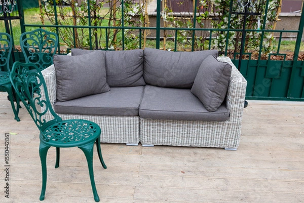 Fototapeta A modern grey sofa with cushions is installed in a glass greenhouse outside. Lush green plants and vibrant flowers surround the living area, creating a serene atmosphere.